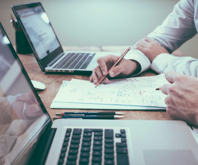 Zwei Hände arbeiten an einem Dokument, während zwei Laptops im Hintergrund stehen.