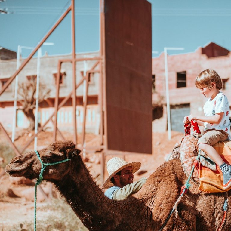 Ein Kind reitet auf einem Kamel, während ein Erwachsener daneben steht. Wüstenlandschaft im Hintergrund.